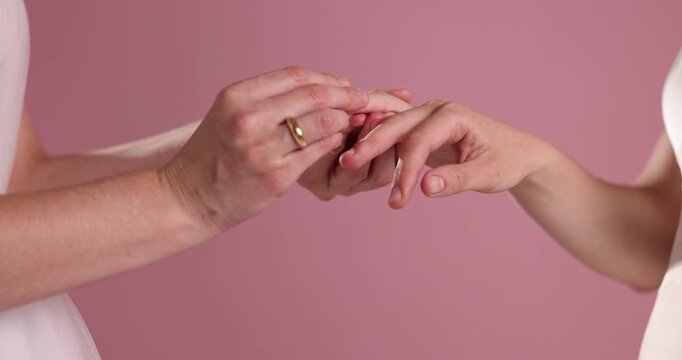 Lesbian wedding. Women putting rings on each others fingers against pink background, closeup
