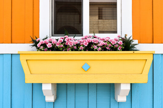 Charming yellow window box planter filled with blooming pink flowers on a vibrant colorful house facade with orange and light blue wooden siding, rustic cottage decor concept.