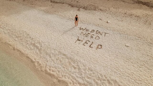 Woman walking on remote Kefalonia beach writing message we dont need help on white pebble shore