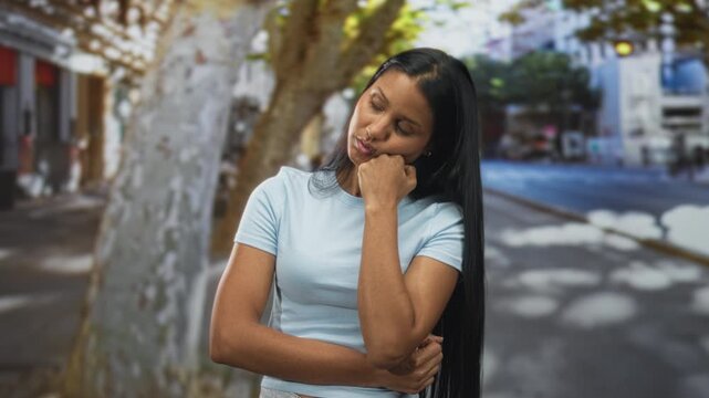 Young hispanic woman resting chin on fist with arms crossed on a city street under plane trees and storefronts; introspection.