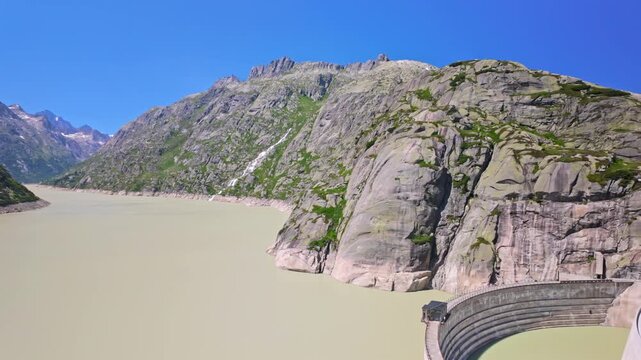 Mountain reservoir of Grimsel pass with a curved concrete dam, pale green water and steep rocky cliffs under a clear blue sky, showing alpine landscape, engineering and remote summer travel.