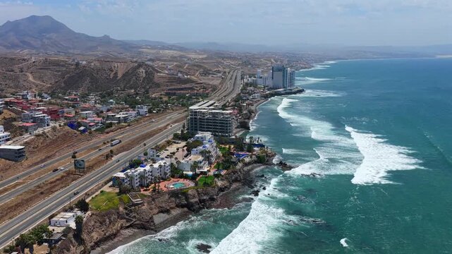 Dolly in aerial toward Las Rocas Resort and Spa along the Rosarito coastline near the Christ of the Sacred Heart monument in Baja California, Mexico with Pacific Ocean waves breaking on rocky shores