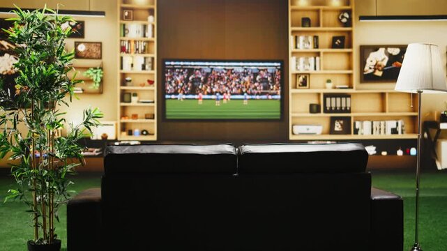 Soccer game is broadcast on TV in an empty apartment living room before fans arrive, with a cozy sofa, plant and screen creating a comfortable home viewing setup.