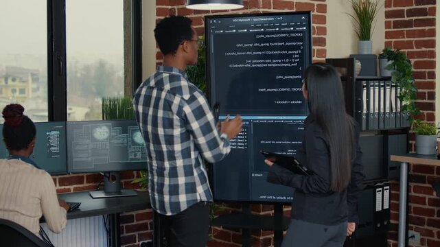 Software developer studies wall screen code during a debugging session with a digital tablet, linking error checking, while handling code diagnostics.