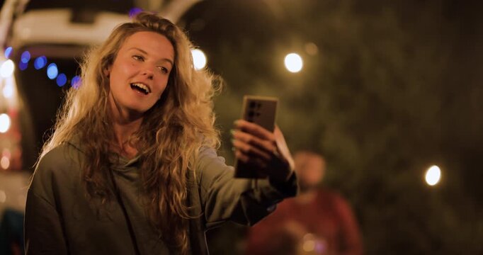 Enjoying a night with friends and taking selfies at a campsite under string lights in nature while having fun and sharing moments together