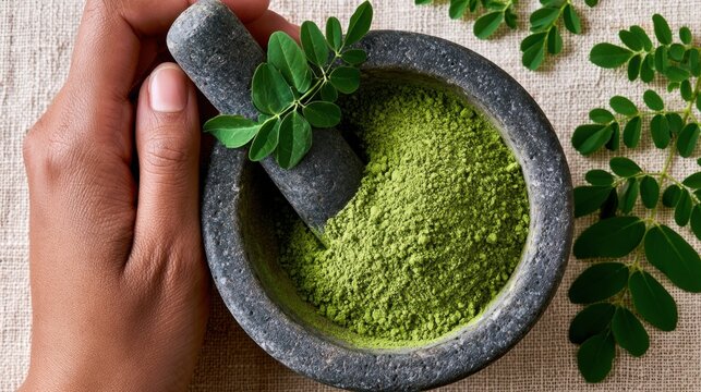 Hand holding stone pestle grinding fresh green moringa leaves into vibrant powder inside stone mortar on rustic fabric background with scattered leaf branches