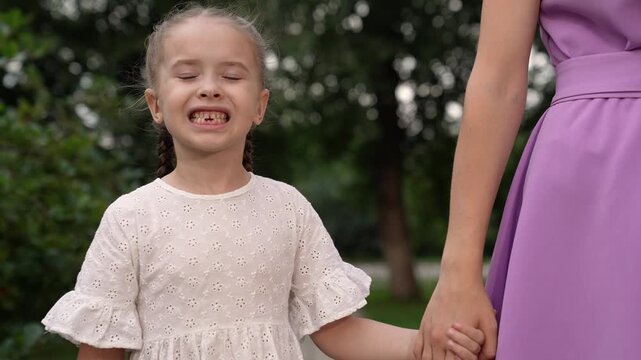 Pretty little daughter without milk teeth on lower jaw smiles holding mother hand in park closeup. Cute daughter shows healthy teeth and holds mother hand in park. Mother and daughter walk in park