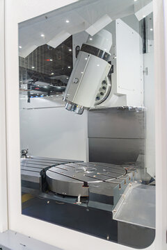 A close-up shot through protective glass of a modern 5-axis CNC milling machine chamber. heavy-duty rotary table, precise tilt-head spindle, and metal working area, advanced industrial manufacturing