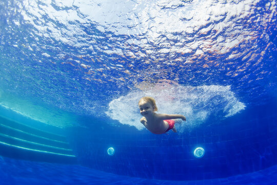 Underwater shot of happy smiling young child jumping with splashes and diving with fun in blue swimming pool. Healthy family lifestyle, water sport activity, swimming lessons on holidays with kids