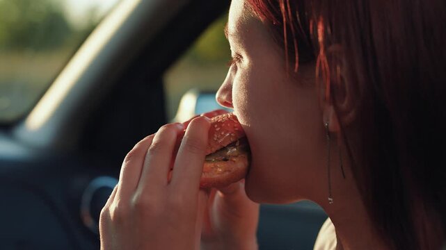 Woman eats delicious burger sitting in car closeup. Woman passenger has nutritious snack for lunch in driving car. Young woman bites burger sitting on passenger seat in driving car at road trip