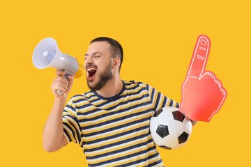 Obraz premium Male sports fan with soccer ball and foam hand shouting into megaphone on yellow background
