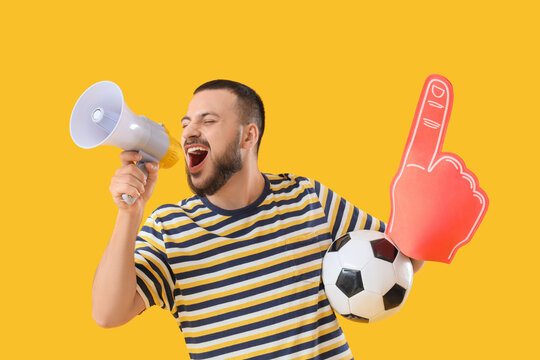 Male sports fan with soccer ball and foam hand shouting into megaphone on yellow background