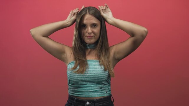 Woman making antlers hand gesture while wearing teal tube top and choker in studio midshot with pink backdrop; playful energy.