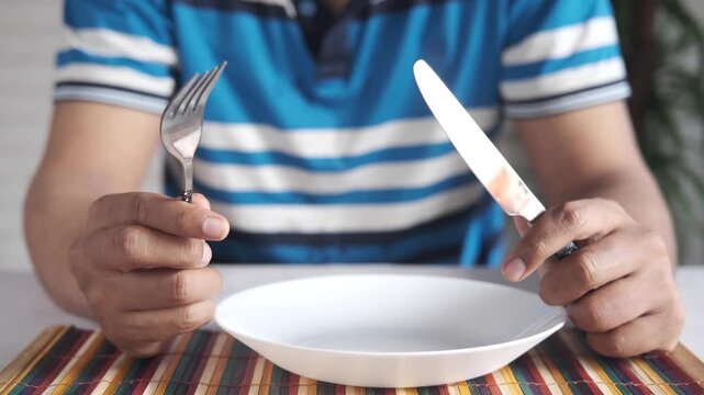 Man holding fork and knife over empty white plate on dining table
