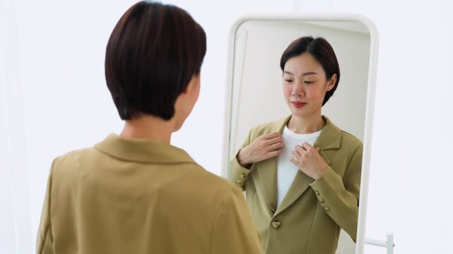 Confident Asian female getting dressed in front of a mirror, preparing for a workday or job interview. Concept of elegant wardrobe selection and modern lifestyle.