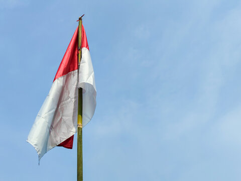 Indonesian Red and White Flag on a Bamboo Pole Against a Background of a Blue Sky with Soft Clouds. Suitable for Independence Day Greeting, Featuring a Negative Space on The Right Side.