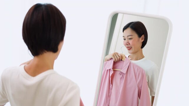 Cheerful Asian female holding hangers with casual shirts, preparing her outfit in a mirror. Concept of personal style development, dressing up and modern lifestyle.