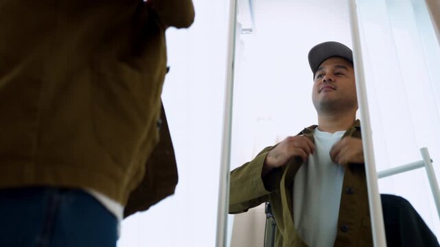 Smiling Asian man in a baseball cap looking in a mirror, adjusting his brown casual shirt jacket. Concept of morning preparation, urban street fashion and everyday confidence.