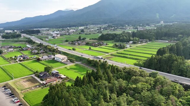 Aerial view captures a quiet rural town nestled among green fields. Mountains loom in the distance, framing a peaceful countryside scene. Roads cut through fields, connecting homes