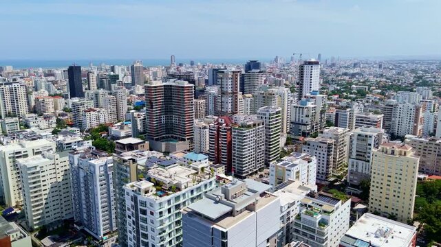 Dense high-rise condominiums fill sprawling urban skyline in Santo Domingo, Dominican Republic, rooftop pools plus balconies stretch toward distant Caribbean coastline. Aerial drone wide shot