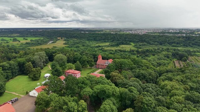 Drone video showcases Raudondvaris Castle surrounded by dense green forest, formal gardens, historical redbrick buildings, and dramatic skies. Bright daylight, wide angle, scenic aerial footage.