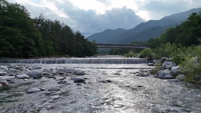 Rivers rush over rocks, reflecting clouds above. Trees line banks, framing distant mountains. Bridge spans water, blending nature with human presence. Peaceful flow suggests quiet, natural beauty