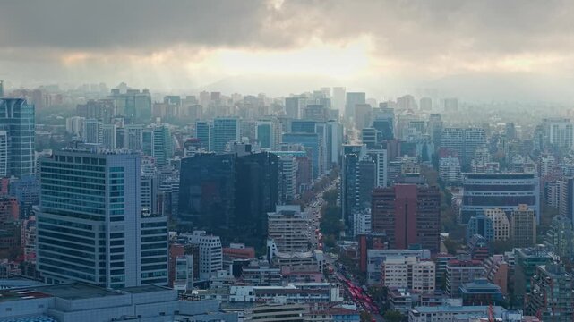Telephoto aerial orbit of modern buildings in Santiago, Chile, with the sun in the background and the city beneath a large gray cloud
