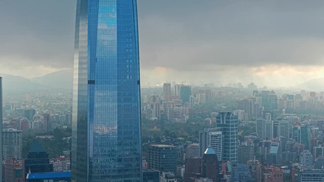 Bird's-eye view ascending from Santiago, Chile, with the modern Costanera Tower and a large gray cloud over the city, with the sun in the background