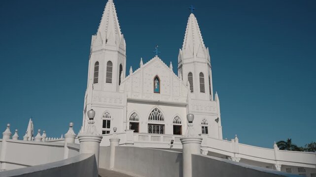 Wide shot of Velankanni Basilica twin white Gothic spires and blue crosses standing against a blue sky, ornate lamp posts framing the historic Catholic church. Basilica of Our Lady of Good Health.