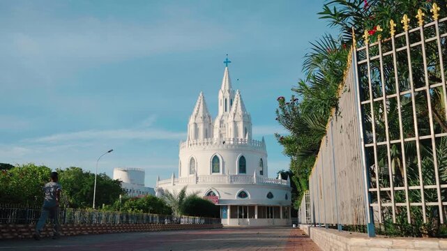 Time lapse of clouds drifting over the rounded white Gothic tower of Velankanni Basilica, palm trees and an ornate gate lining the quiet courtyard under a clear blue sky, Tamil Nadu, India