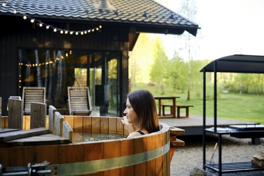 A woman sits in a rustic wooden hot tub surrounded by greenery at a countryside retreat. The setting features a tiny house with string lights, promoting a tranquil atmosphere