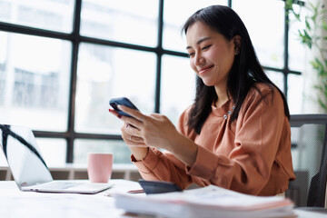 Obraz premium Young asian businesswoman sitting at office desk, messaging on smartphone while working, managing tasks, using technology for communication and remote work