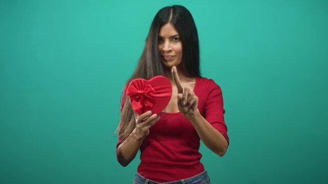 Woman holding red heart gift box points index finger in studio, wearing red top and jeans with teal backdrop; playful caution.