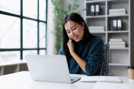 Young asian woman working remotely on laptop and smartphone, engaging in a business call while smiling at her home office desk, focusing on communication and connectivity