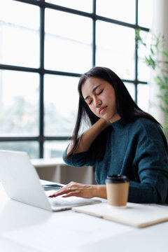 Young woman experiencing neck pain and discomfort from sedentary work while using a laptop in a modern office, feeling tired, stressed, and overworked