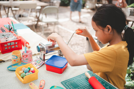 Focused Asian kid using a metal rolling machine to flatten polymer clay for art crafts on an outdoor table