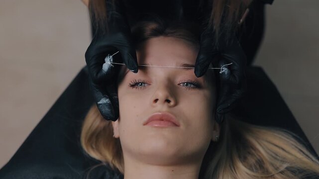 eyebrow threading, beauty salon, black gloves. Close-up beautician wearing black nitrile gloves performing eyebrow threading on young woman in professional beauty salon, highlighting precision