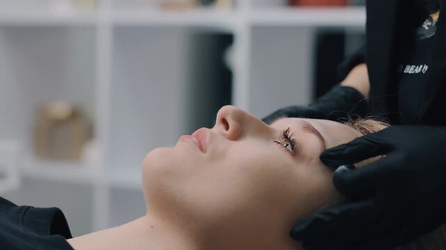 beauty treatment, professional beauty, salon hygiene. Beauty specialist wearing black nitrile gloves carefully performing eyebrow threading for young female client in clean professional salon