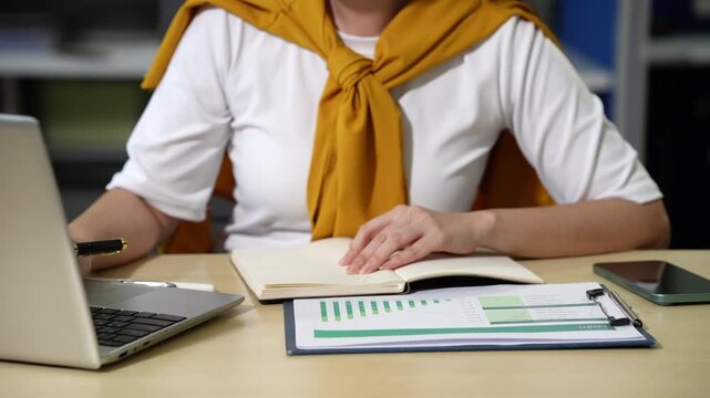 Person working at a desk with laptop, notebook, documents, and smart phone, taking notes and reviewing paperwork in a home office or study setting.