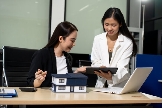 Two businesswomen discussing a house model while reviewing data on a tablet in a modern office, with a laptop on the desk