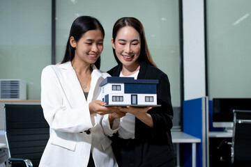 Obraz premium Two smiling businesswomen holding a model house in an office, showing a real estate concept, teamwork, and property investment