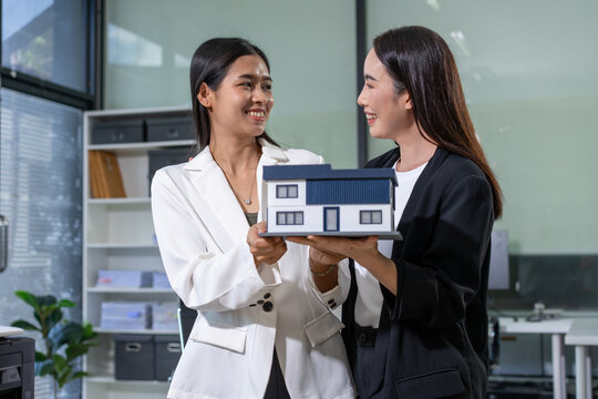 Two businesswomen smiling while holding a small house model in an office, suggesting real estate planning, property sales, and teamwork