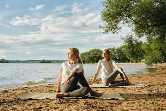 Two women practice yoga on a sandy lakeshore, surrounded by the calm waters and lush greenery. The sun illuminates their peaceful faces as they embrace the tranquility of nature in their activity