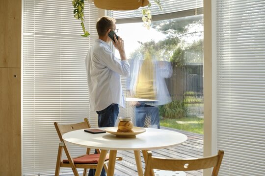 A middle-aged man stands near a large window, engaged in a phone conversation. Sunlight filters through the blinds, illuminating a contemporary dining area with a circular table and simple decor