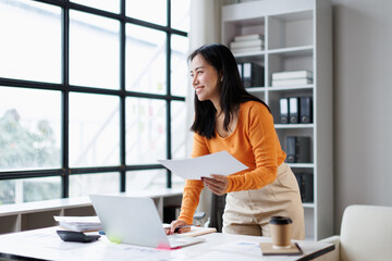 Asian businesswoman smiling, standing at her office desk, holding paperwork, analyzing reports, and...