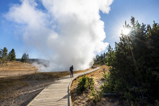 Hiker on footbridge next to steaming hot spring, sun with sun star, Churning Caldron, Yellowstone National Park, Wyoming, USA