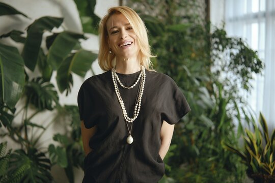 A cheerful woman with blonde hair poses confidently amidst a backdrop of vibrant plants in a bright indoor area. She wears a stylish outfit with layered necklaces, exuding a sense of warmth and joy