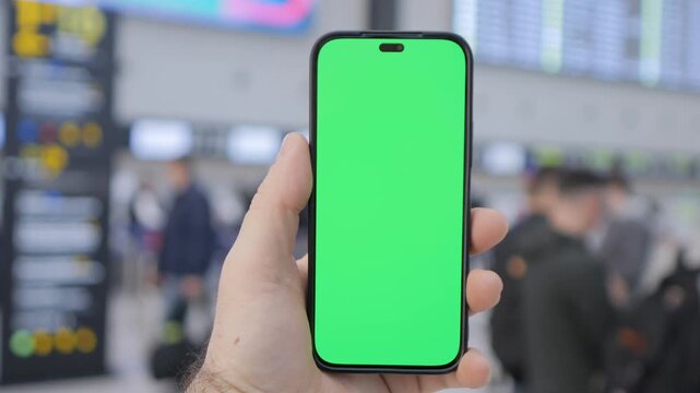 Hand holds phone with green screen at busy airport. Background shows travelers and digital boards. Perfect for app demos or travel-related ads