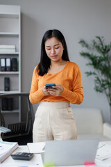 Fototapeta premium Young asian woman standing in a modern office. Engrossed in her smartphone. Managing business communications and tasks