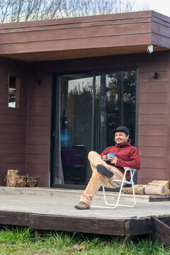 Man crossing his legs and smiling with a hot drink on a deck outside a modern wooden cabin